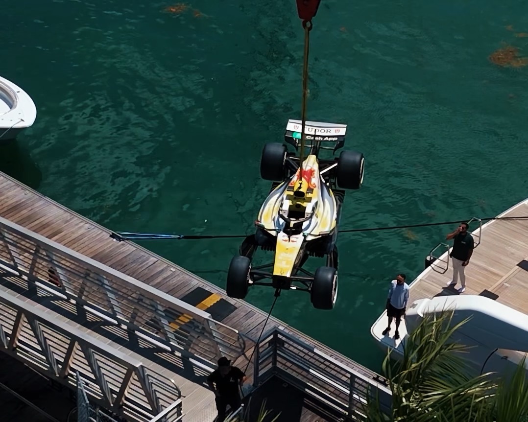 A Formula 1 car suspended by crane above the water beside a Maritime yacht